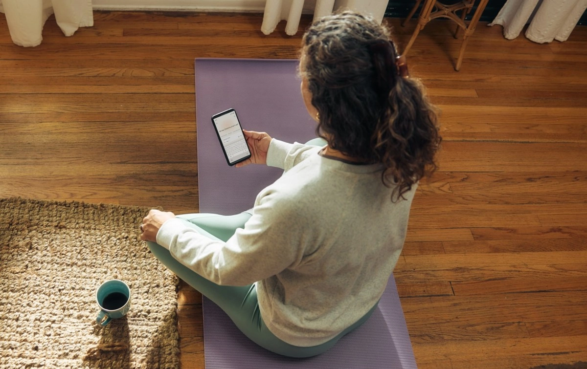 Woman uses phone on yoga mat with coffee.